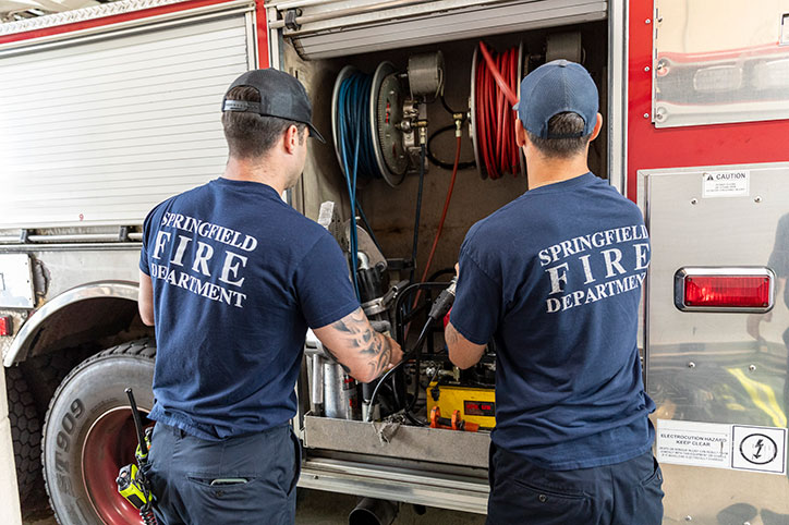 Two firefighters from the Springfield Fire and Emergency Serivces working with equipment in a compartment on their fire truck.