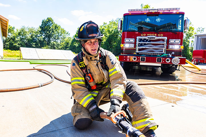 A firefighter in turnout gear kneeling on the ground holding a fire hose in front of a Pierce fire truck.