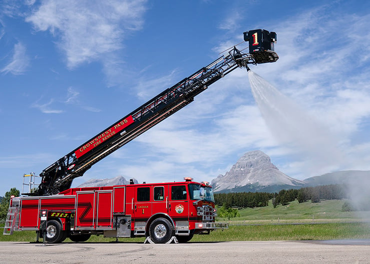 The officer's side of Crowsnest Pass Fire Rescue's Enforcer 110' Ascendant Heavy-Duty Platform with the aerial extended spraying water in front of a mountain.