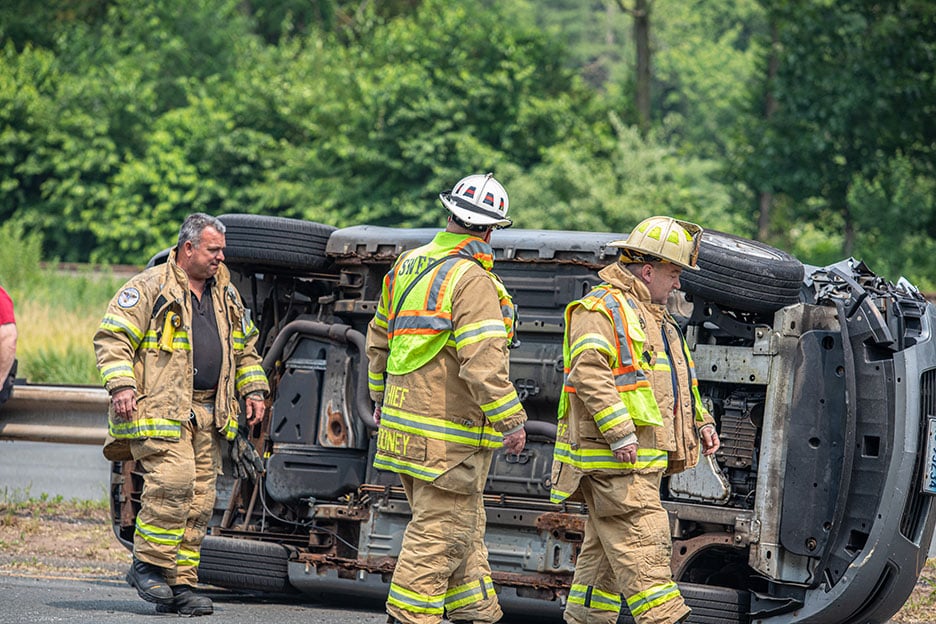 Three firefighters in turnout gear walking on scene of an over-turned vehicle on a road.