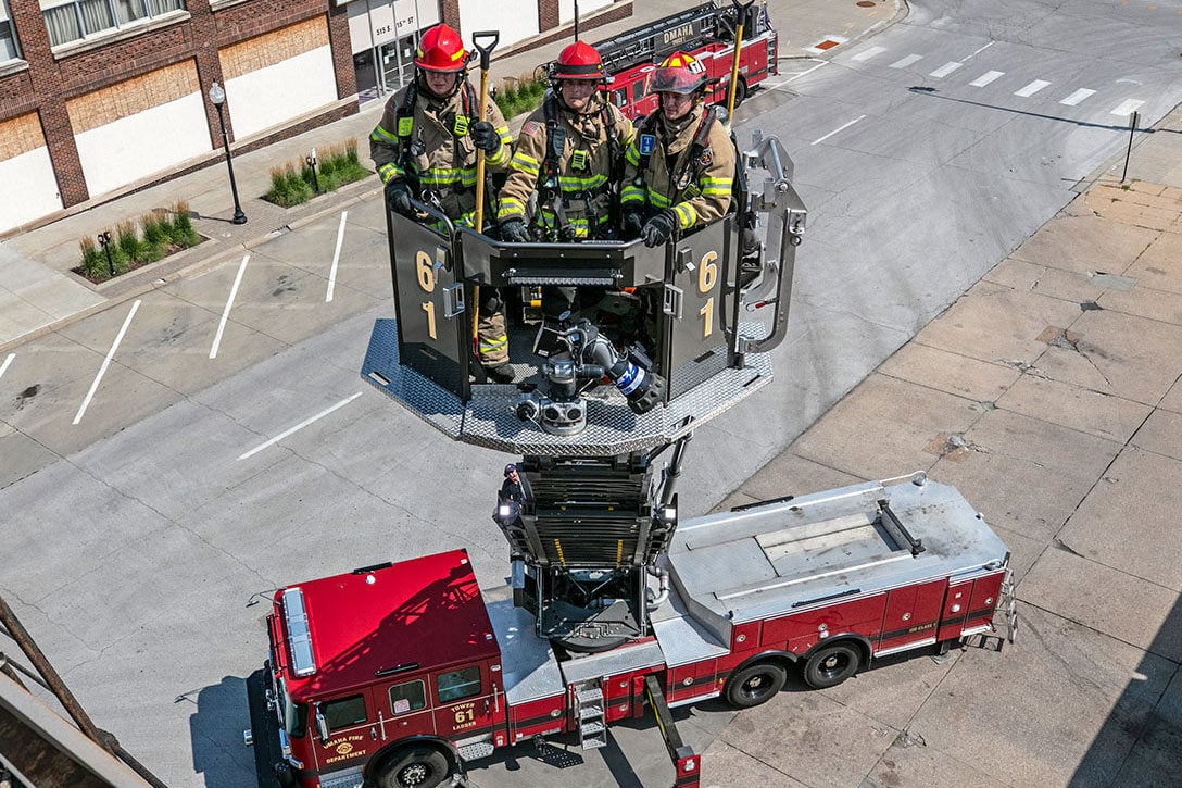 Three firefighters in turnout gear in a bucket of an Ascendant Aerial Tower next to a rooftop with tools in their hands.