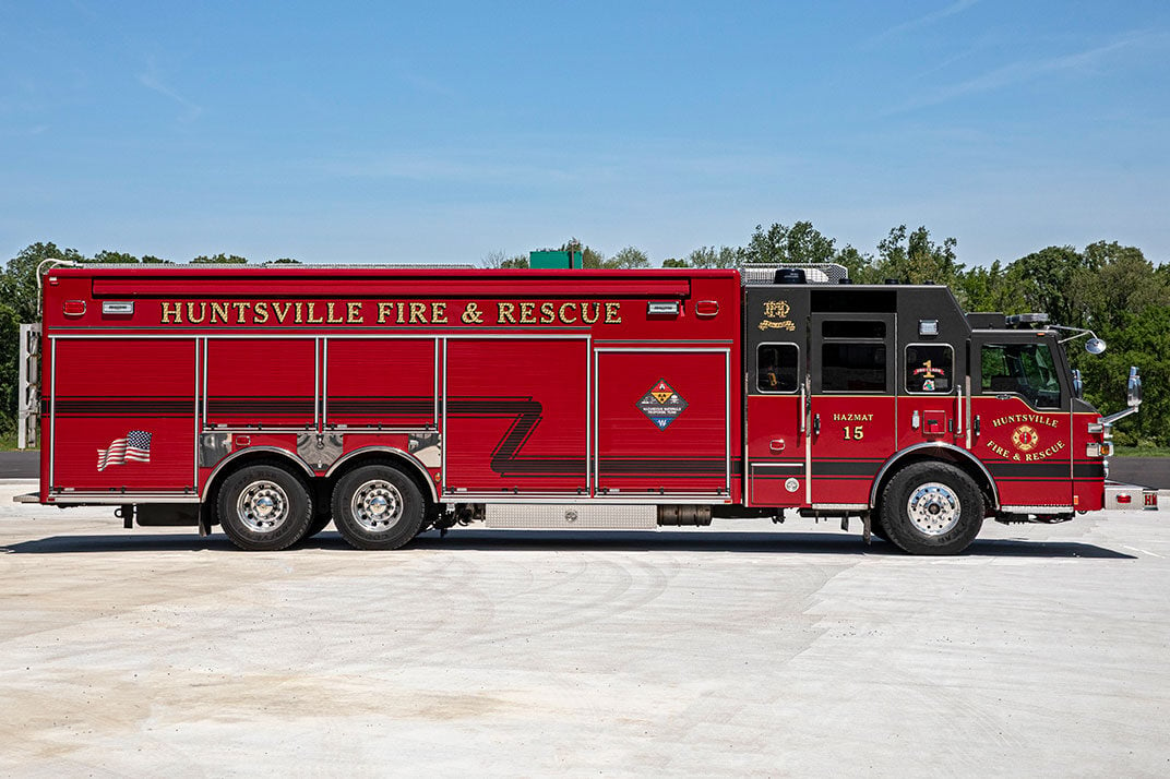 The officers side of a combination heavy-duty rescue fire truck on a sunny day parked in front of trees.