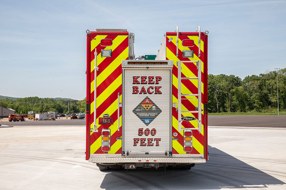 The rear of a heavy-duty rescue fire truck in a parking lot surrounded by trees on a sunny day.