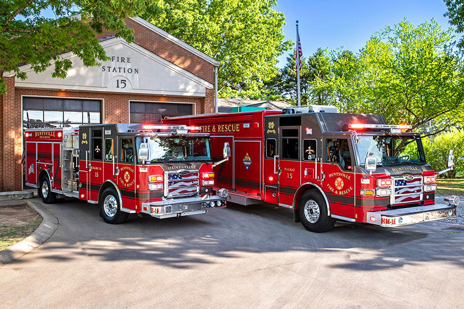 Two fire trucks parked in front of a brick fire station and trees on a sunny day.