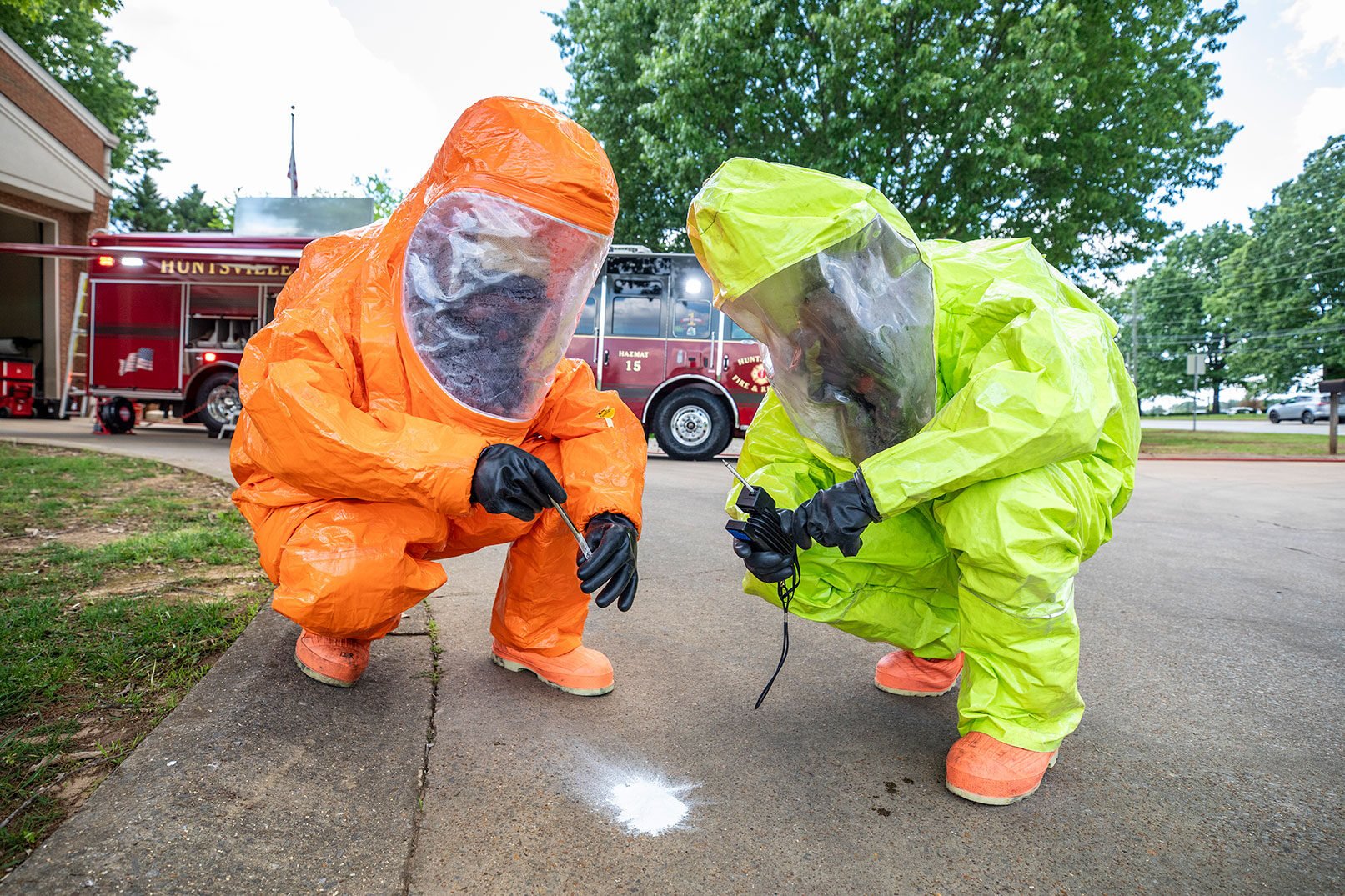 Two firefighters in neon hazardous material suits looking at a white substance on the ground in front of a fire truck.