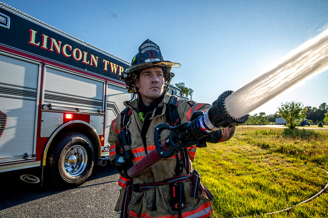 A firefighter in turnout gear holding a fire hose in front of a fire truck on a sunny day.