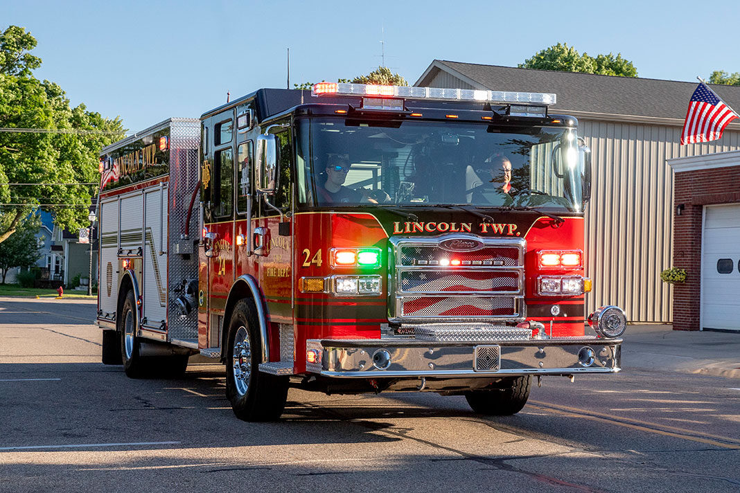 An Enforcer Heavy-Duty Rescue Pumper driving on a street in front of a fire station with the lights on.