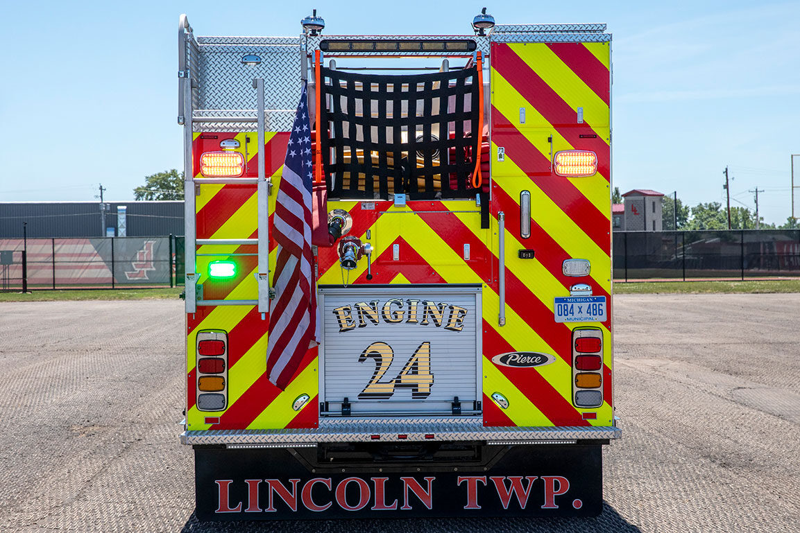 The rear of a Rescue Pumper fire truck with the lights on and an American flag hanging off the rear in a parking lot on a sunny day.