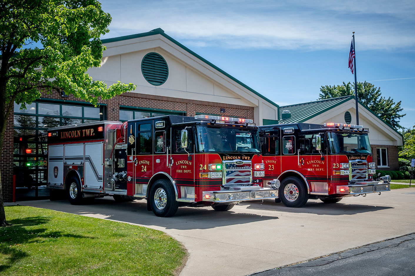 Two fire trucks with their lights on parked in front of a fire station on a sunny day.