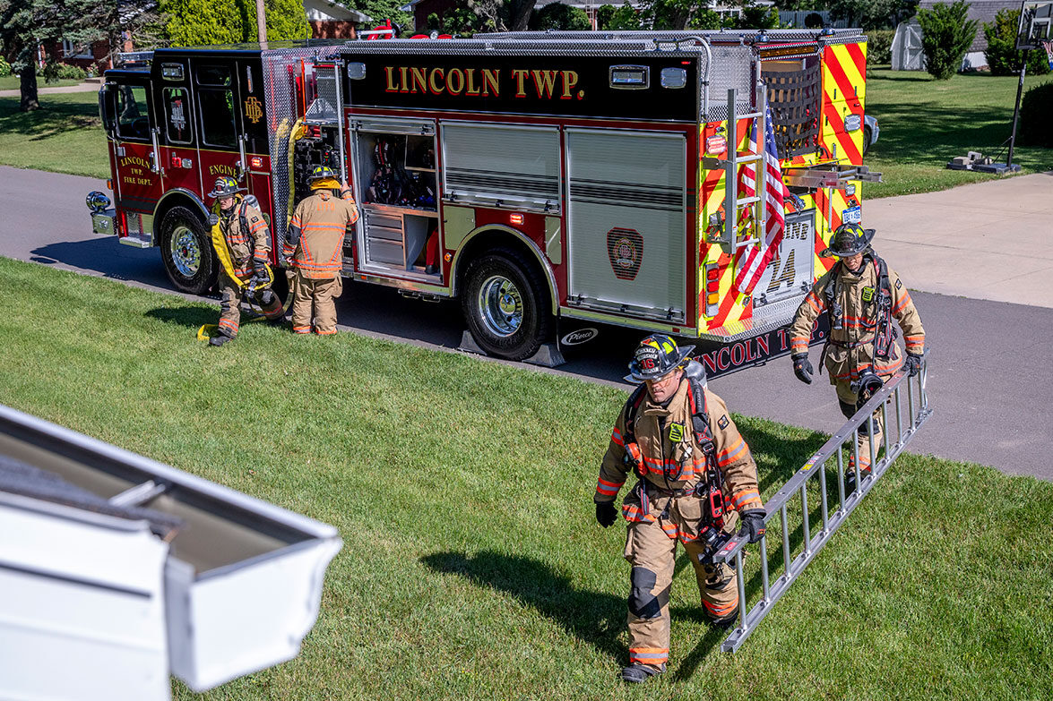 Two firefighters in turnout gear carrying a ladder to a house, a firefighter carrying hose and another standing by the fire truck pump.