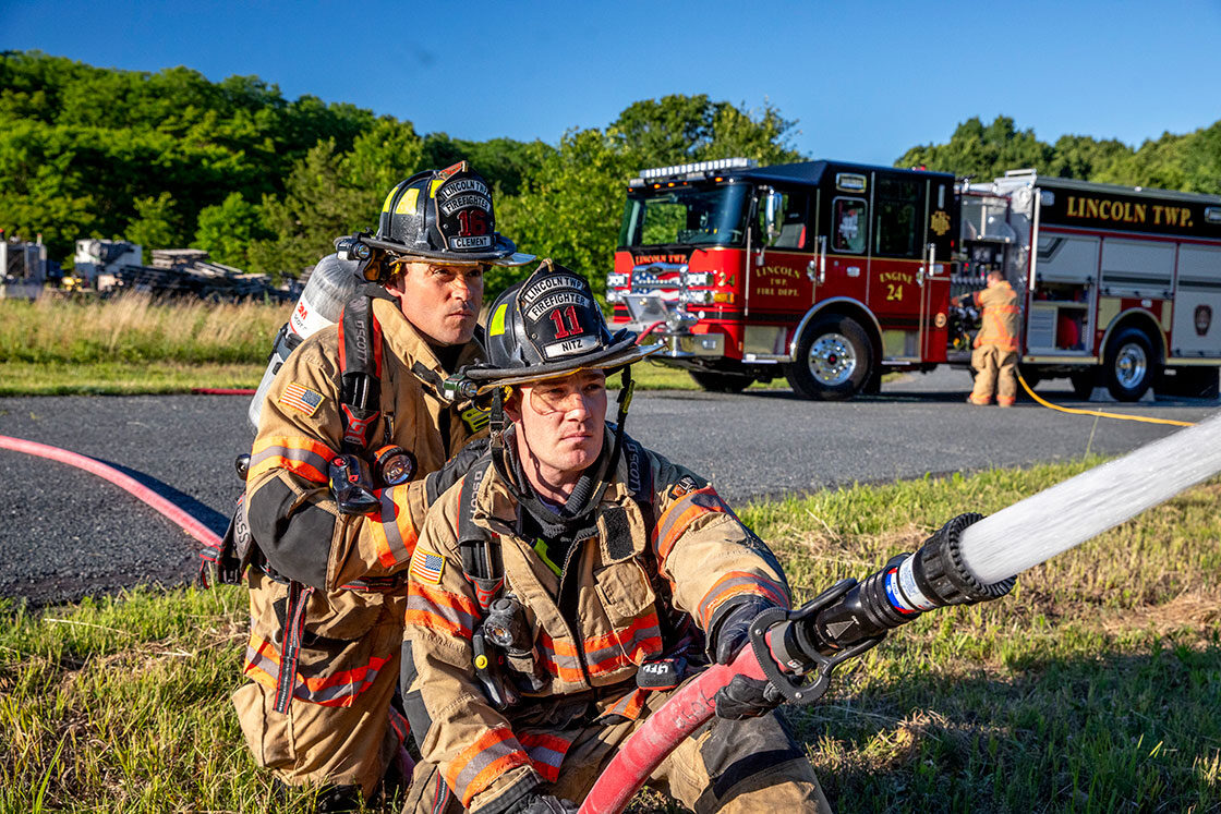 Two firefighters in turnout gear kneeling on the ground holding a fire hose in front of a fire truck with a firefighter at the pump.